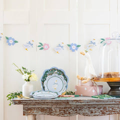 Decorative table with a cake stand, plates, and flowers against a white wall with floral decals.